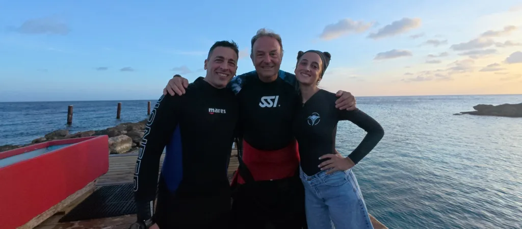 Joe, Leo Tournier y Mile abrazados y sonriendo en el muelle al atardecer, después de bucear