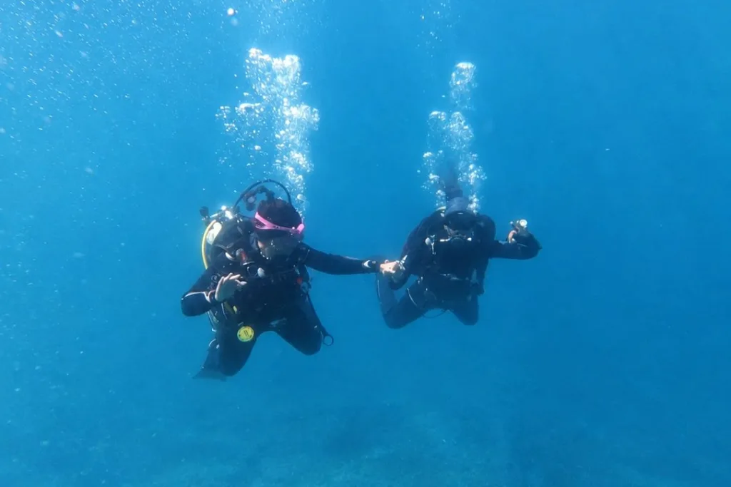 Dos personas buceando de la mano en agua azul, con burbujas subiendo