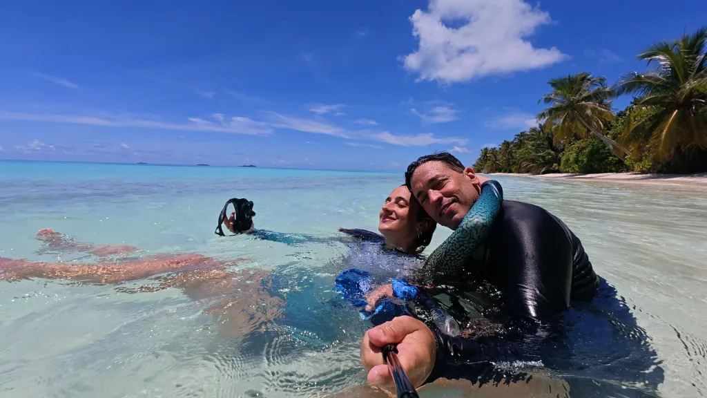 Mile flotando en el agua turquesa y Joe abrazándola, frente a una playa con palmeras en Maldivas